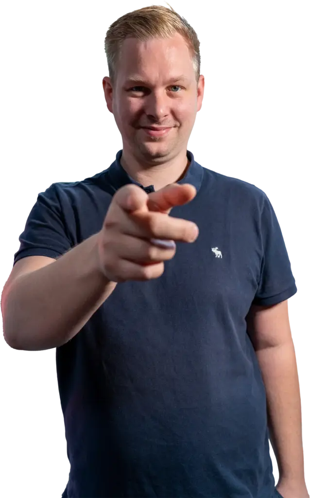 Mann mit blondem Haar in dunkelblauem Poloshirt, lächelnd und auf die Kamera zeigend, Vorderarm prominent im Bild.