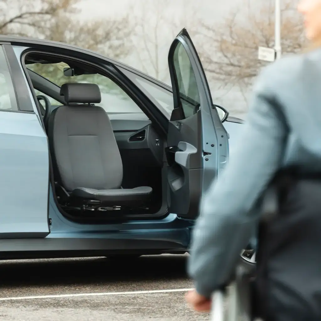 Blauer Pkw mit geöffneter Fahrertür und leerem Vordersitz, unscharfe Person im Rollstuhl im Vordergrund.