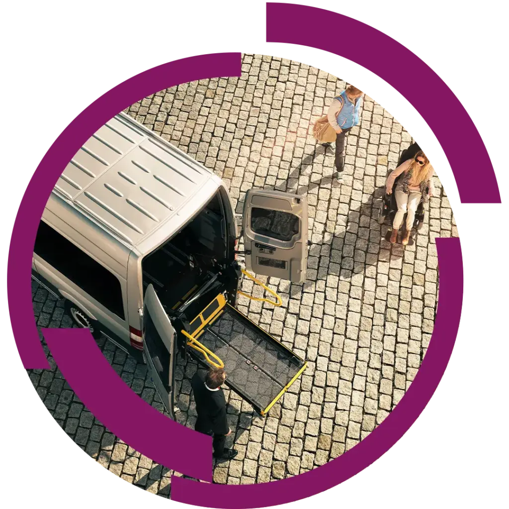 Rollstuhlgerechter Transportbus mit geöffneter Ladebrücke auf Pflaster, daneben Begleitperson und Rollstuhlfahrerin, Blick von oben.