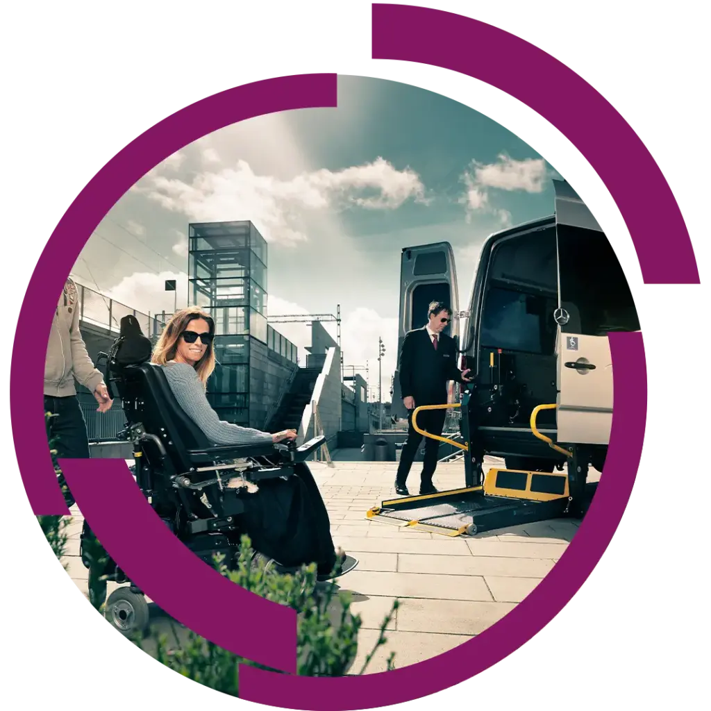 Frau im Elektrorollstuhl lächelt vor einem Kleinbus mit ausklappbarer Rampe, Begleiter und moderner Bahnhofsarchitektur im Hintergrund.