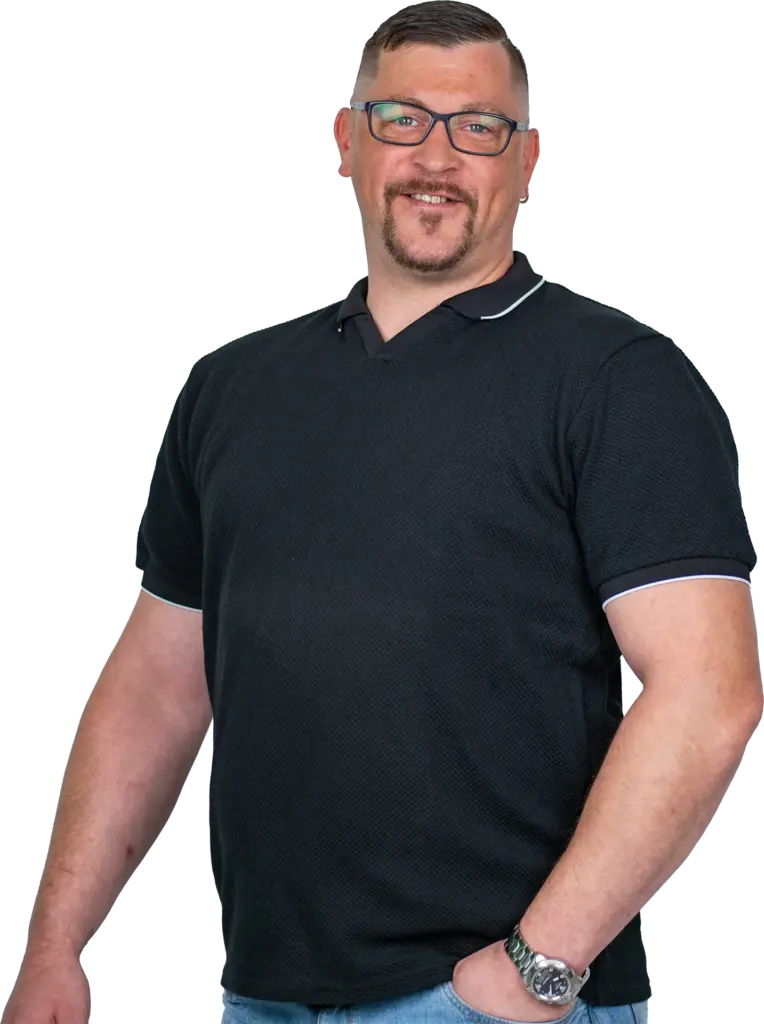 Mann mit Brille, kurzem Haar und Bart lächelnd, trägt schwarzes Poloshirt, eine Uhr und hat eine Hand in der Jeanstasche.