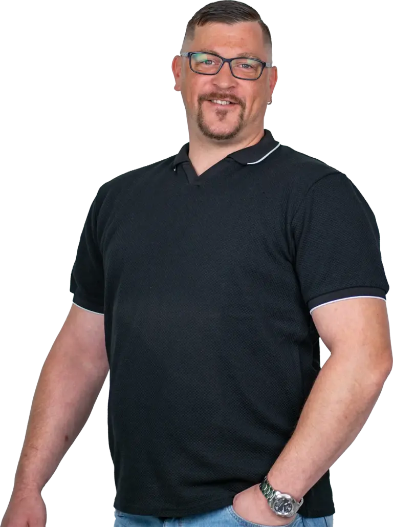 Mann mit Brille, kurzem Haar und Bart lächelnd, trägt schwarzes Poloshirt, eine Uhr und hat eine Hand in der Jeanstasche.
