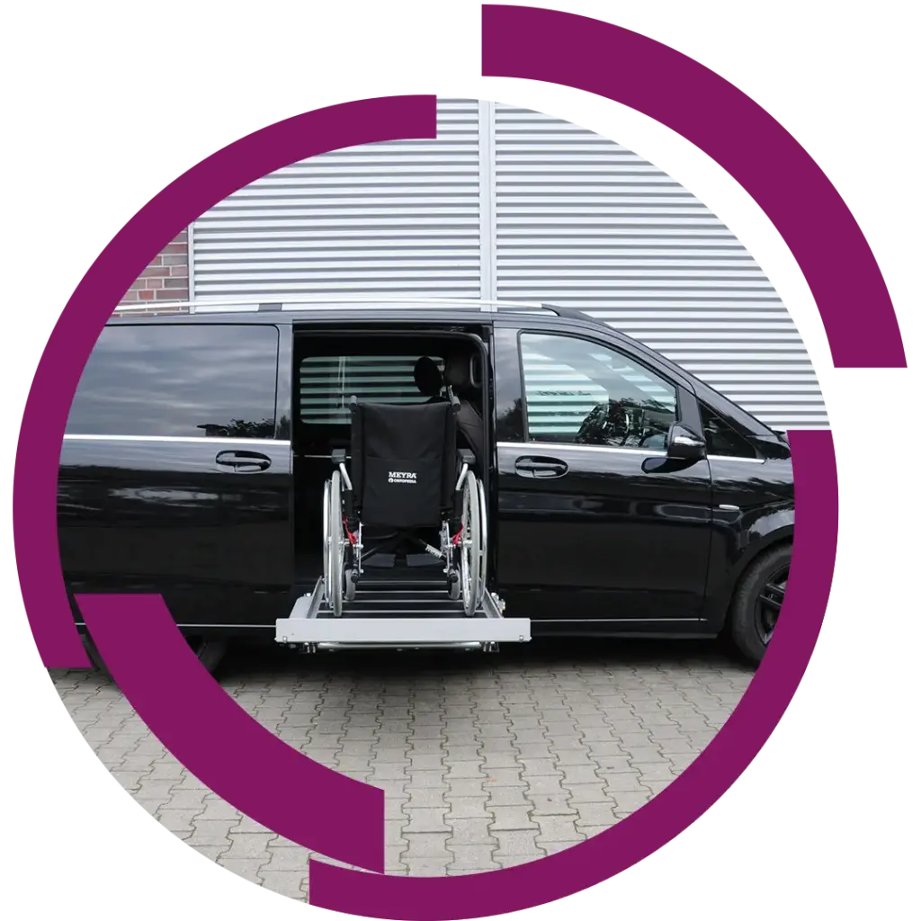 Schwarzer Kleinbus mit seitlich ausgefahrener Hebebühne, darauf ein Rollstuhl, geparkt vor einer Metallfassade.