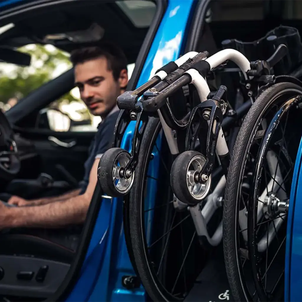 Mann sitzt im blauen Auto, faltbarer Rollstuhl mit kleinen Vorderrädern am geöffneten Türrahmen befestigt.