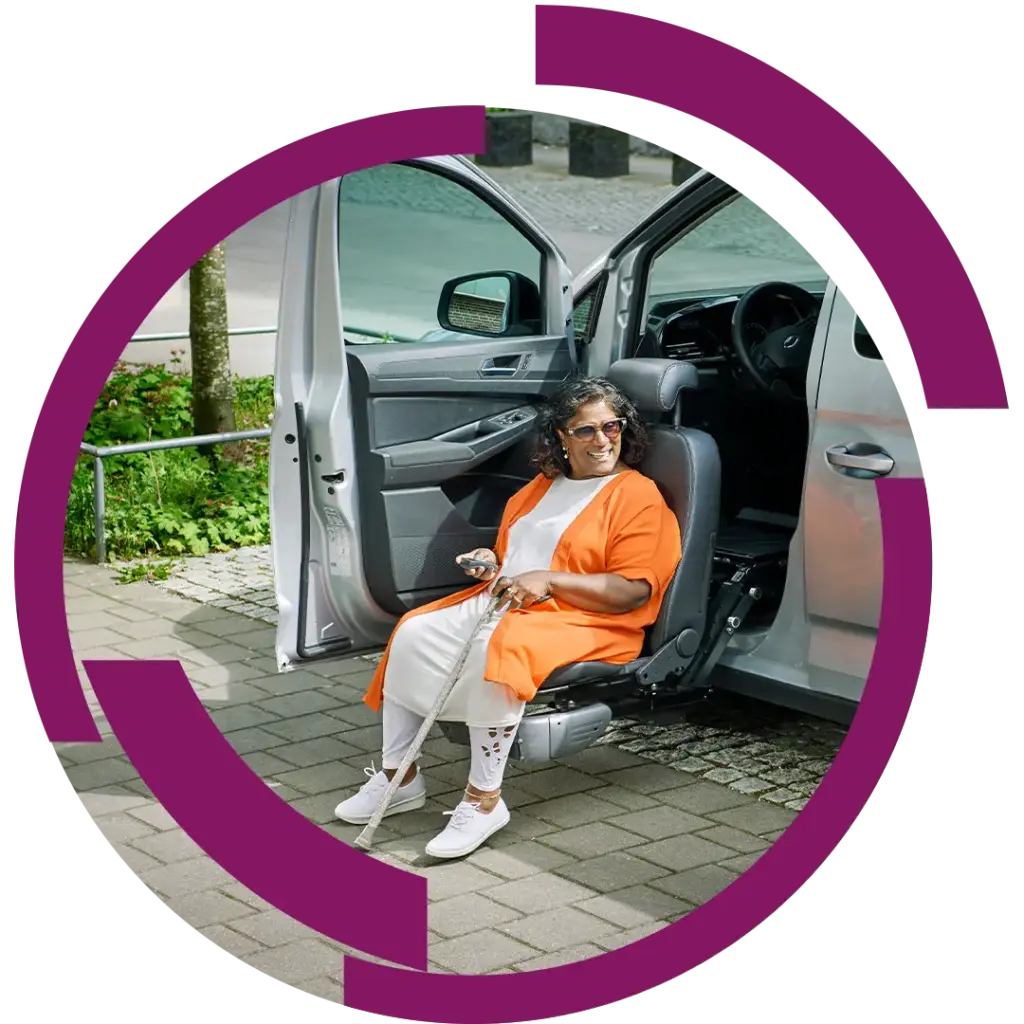 Ältere Frau mit Gehstock steigt lächelnd aus einem Autositzlift, trägt orangefarbene Jacke und weiße Kleidung.