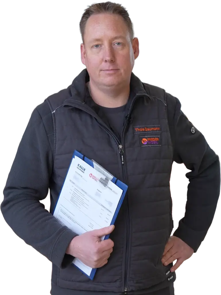 Mann mittleren Alters in dunkler Arbeitsweste mit Namensschild, hält Klemmbrett mit Unterlagen und schaut in die Kamera.