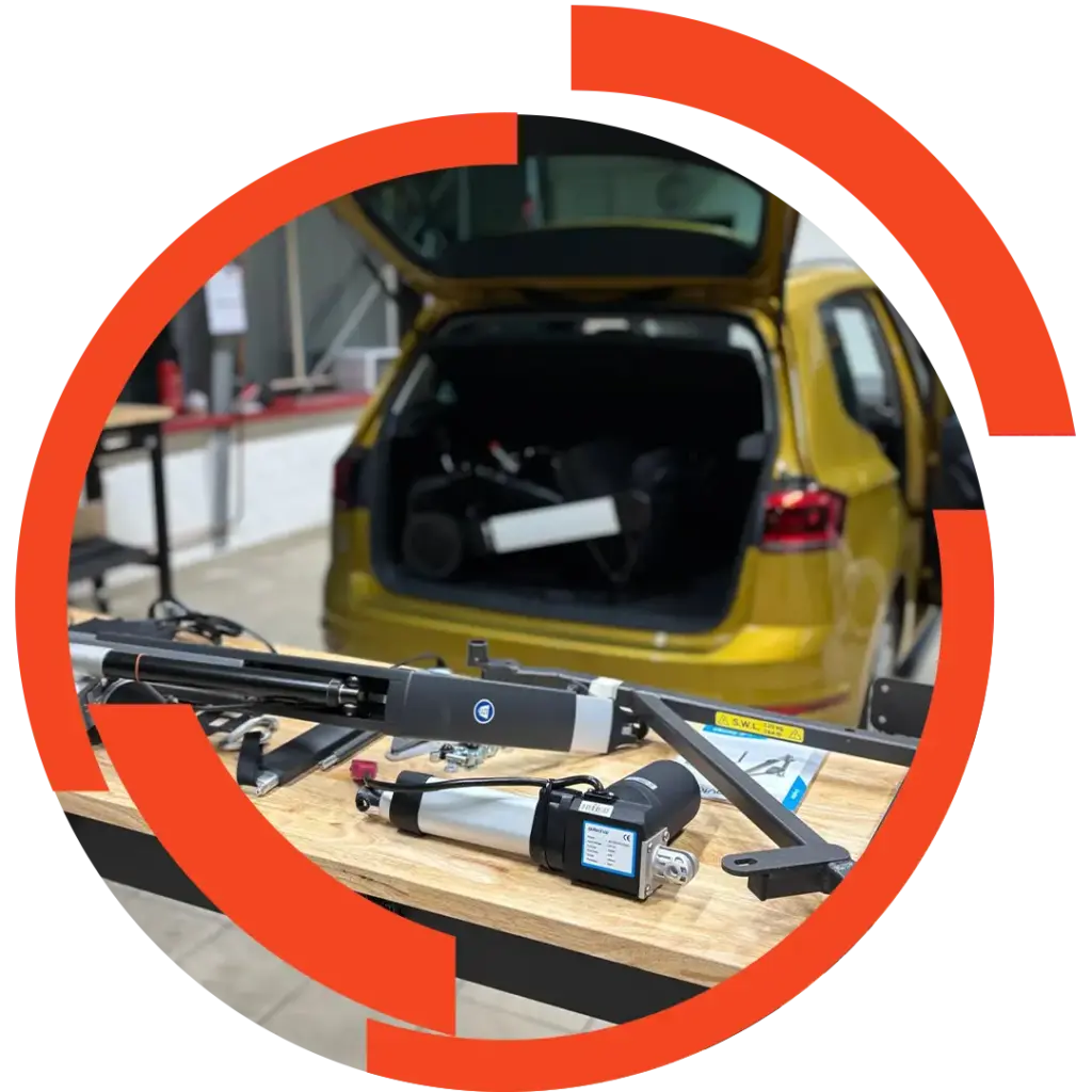 Werkbank mit Elektromotoren und Mechanikteilen im Vordergrund; geöffneter goldener Kofferraum eines Fahrzeugs im Hintergrund.