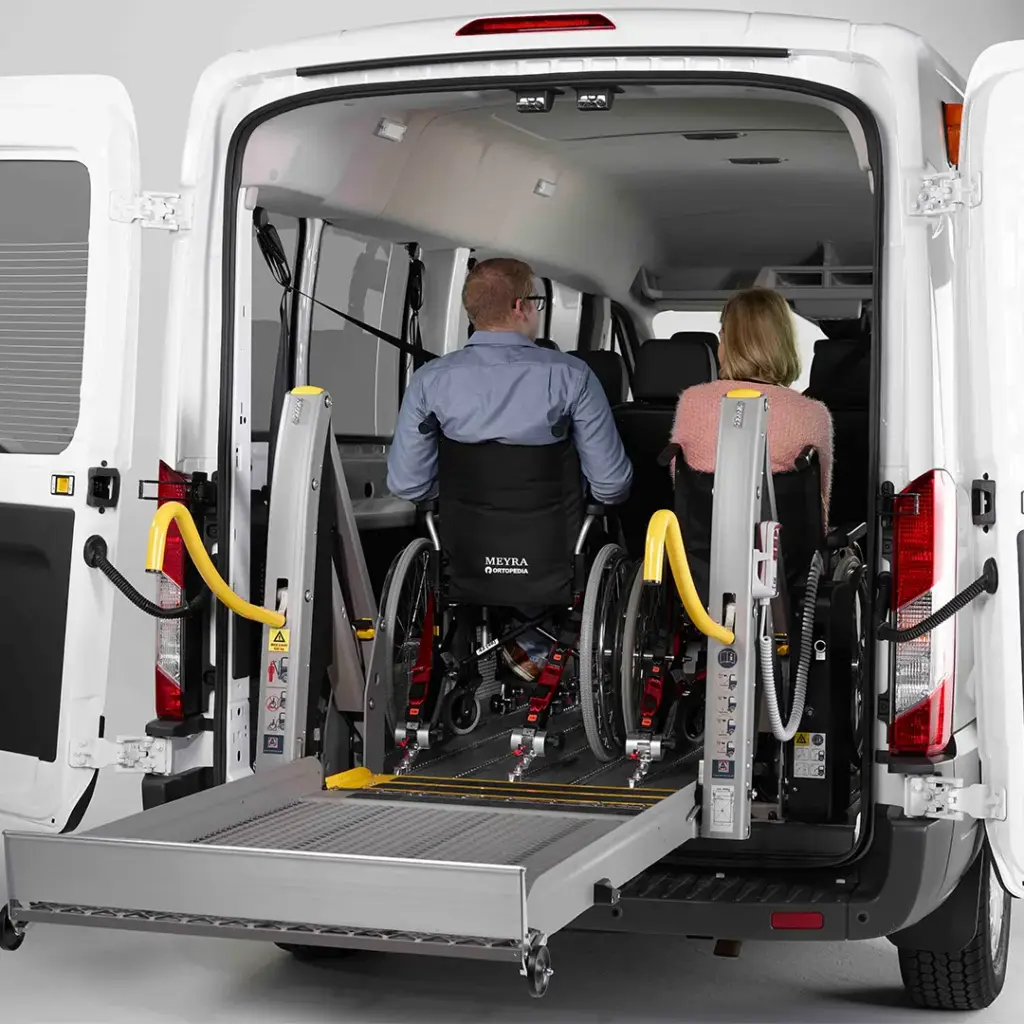 Heckansicht eines weißen Transporters mit geöffneter Ladefläche und Rampe, zwei in Rollstühlen sitzende Personen gesichert auf einer Hebebühne.