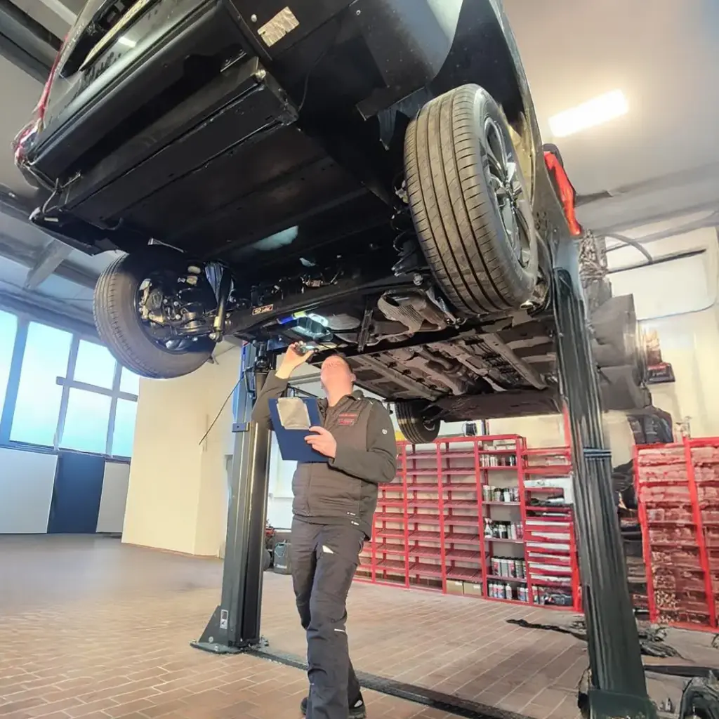 Mechaniker unter gehobenem Auto auf Hebebühne, prüft Unterboden mit Taschenlampe und hält Klemmbrett in einer hellen Werkstatt.