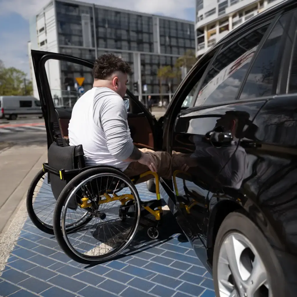 Mann in Rollstuhl schiebt sich seitlich in ein geöffnetes Auto auf einem behindertengerechten Parkplatz in städtischer Umgebung.