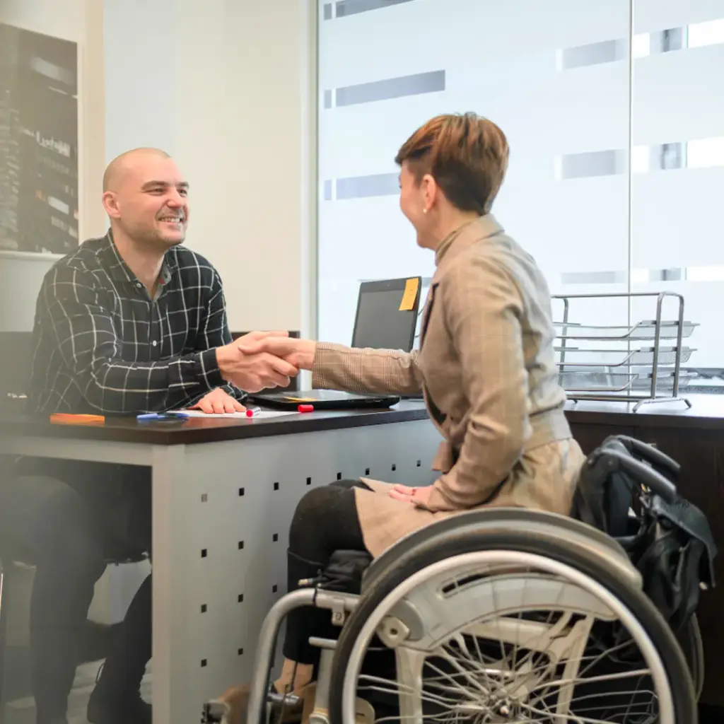 Frau im Rollstuhl schüttelt einem lächelnden Mann am Schreibtisch die Hand in einem hellen Büro.