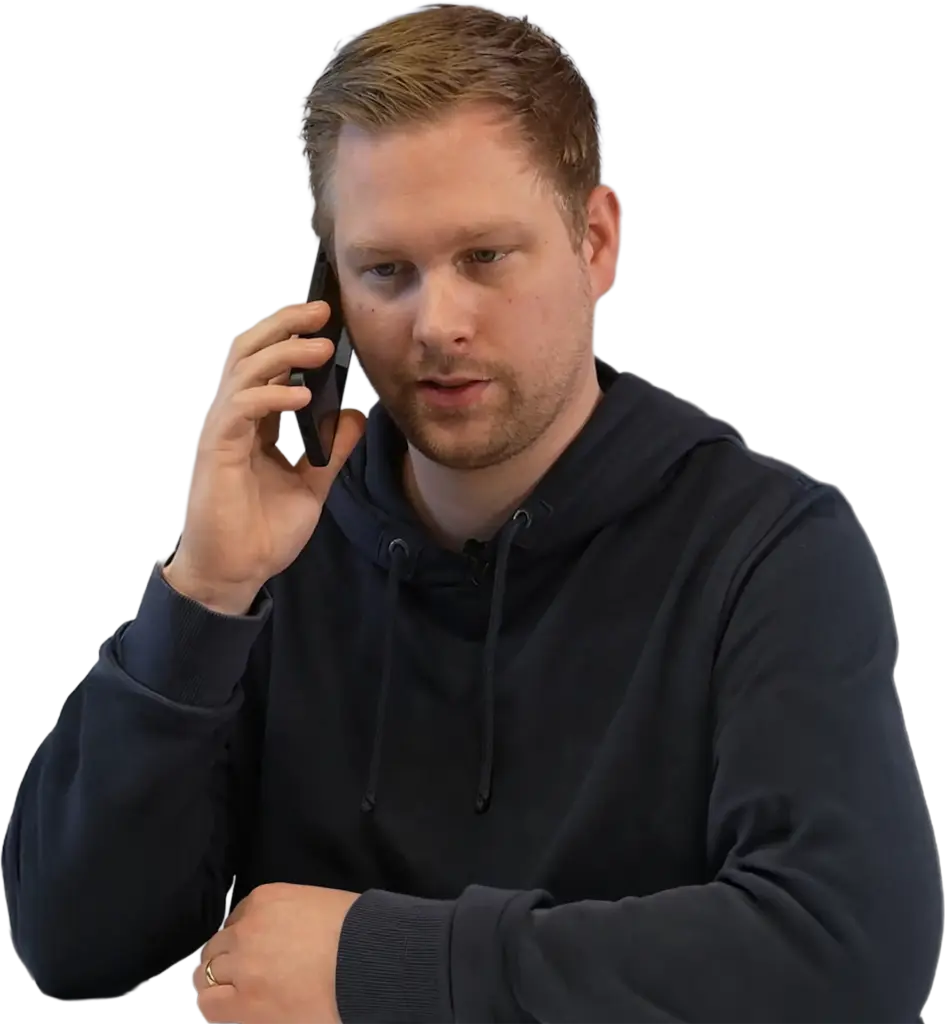 Mann mit kurzem blonden Haar und Bart trägt dunklen Kapuzenpullover und spricht ernst am Smartphone, linker Arm auf dem Tisch, Ehering sichtbar.