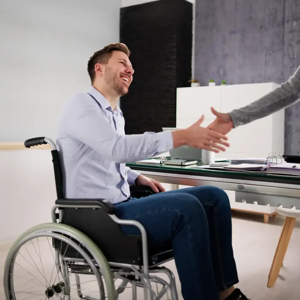 Mann im Rollstuhl sitzt lächelnd an einem Schreibtisch und reicht einer anderen Person die Hand zur Begrüßung.