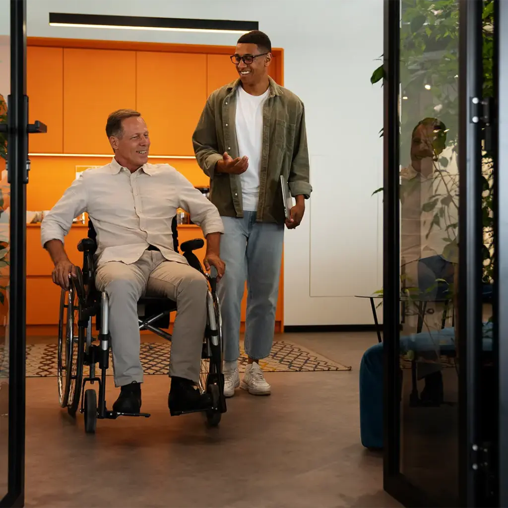 Älterer Mann im Rollstuhl lächelnd, begleitet von einem jüngeren Kollegen mit Laptop, modernes Büro mit orangefarbener Wand im Hintergrund.