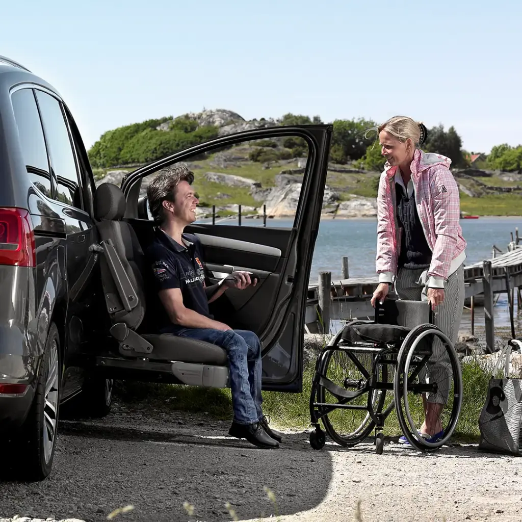Mann sitzt in ausgedrehtem Autositz, lächelnd, während Frau neben einem Rollstuhl am Ufer eines Sees mit Holzsteg steht.