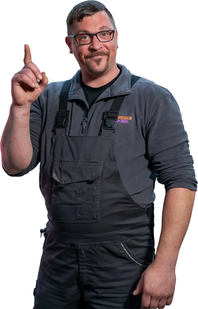 Mann mit Brille und Bart in grauer Arbeitskleidung und Latzhose, lächelnd und mit erhobenem Zeigefinger, vor neutralem Hintergrund.