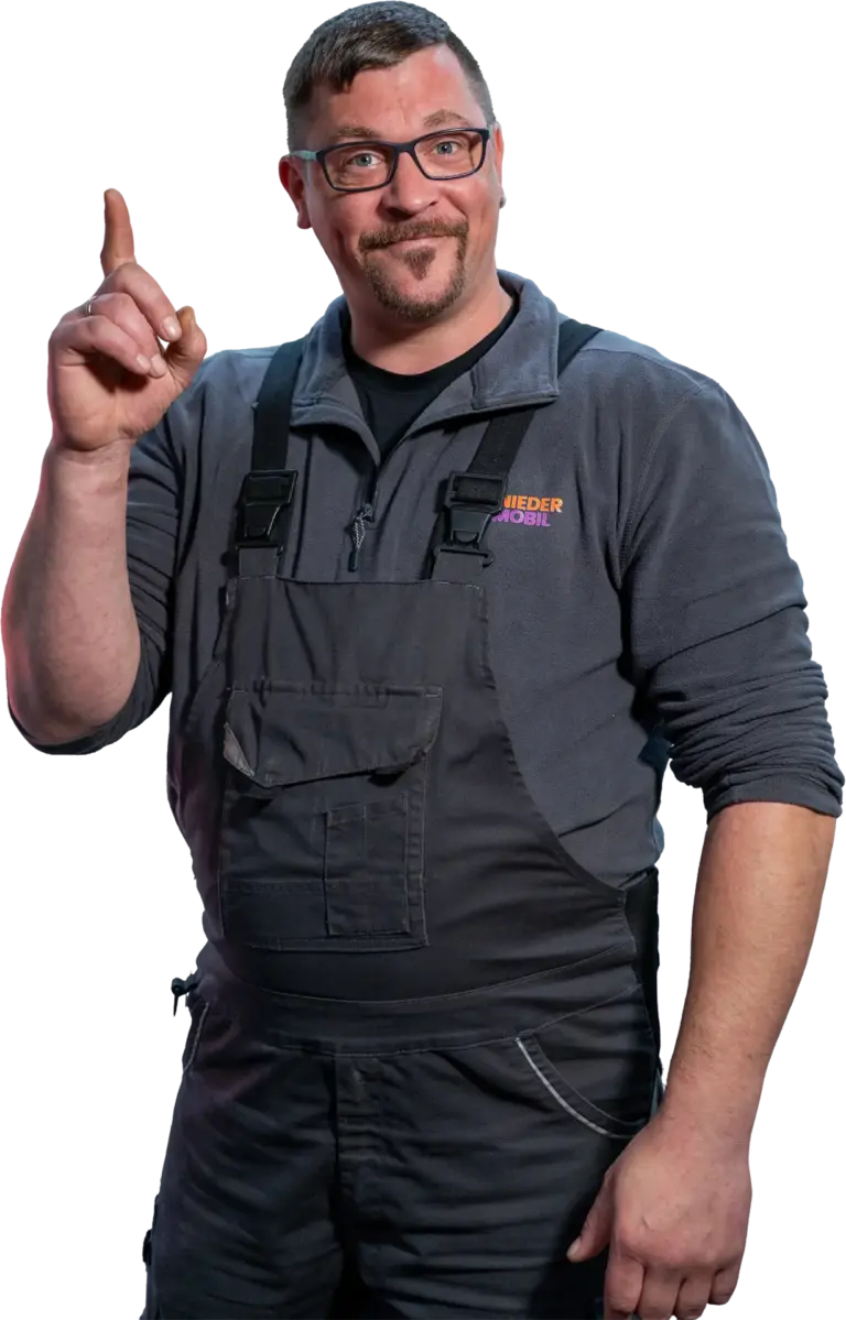 Mann mit Brille und Bart in grauer Arbeitskleidung und Latzhose, lächelnd und mit erhobenem Zeigefinger, vor neutralem Hintergrund.