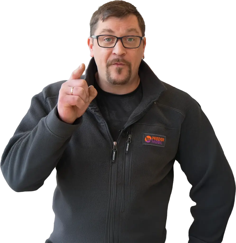 Mann mit Brille und kurzem Bart, zeigt mit erhobenem Zeigefinger und trägt dunkle Fleecejacke mit Wiedermobil-Logo.