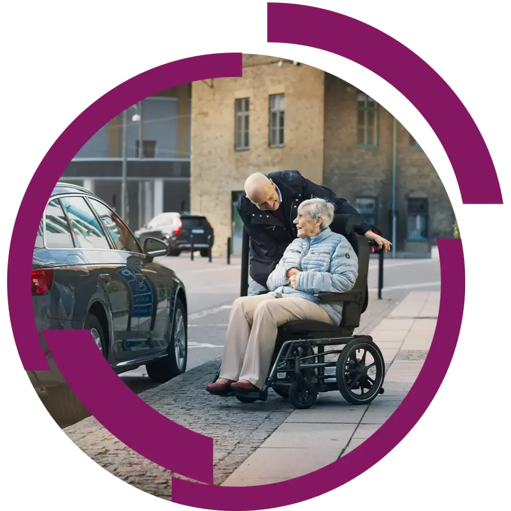 Ältere Frau in Rollstuhl auf Bürgersteig neben geparktem Auto, ein Mann beugt sich fürsorglich zu ihr und spricht mit ihr.