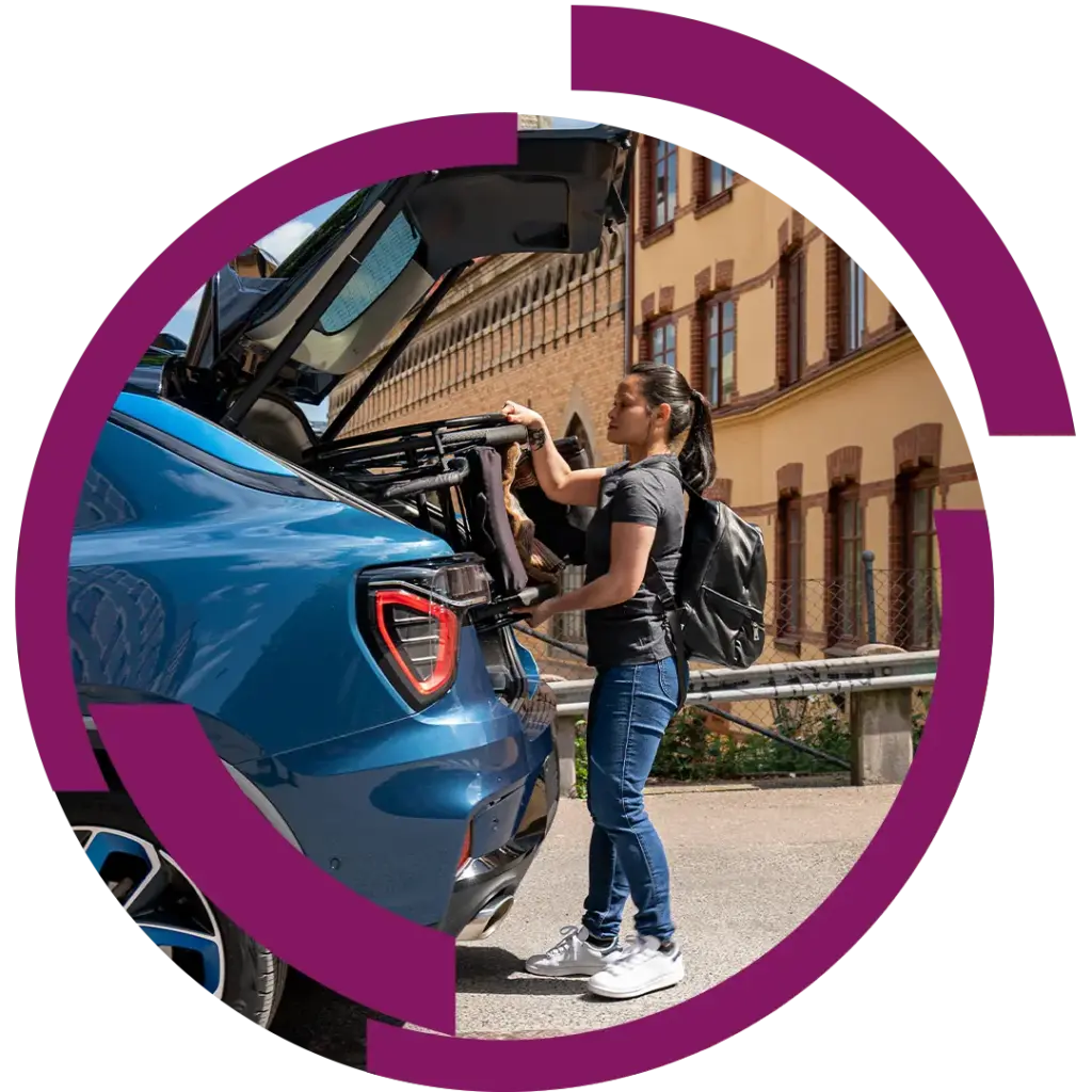 Frau mit schwarzem Rucksack lädt zusammengeklappten Kinderwagen in den Kofferraum eines blauen Autos, Stadthäuser im Hintergrund.