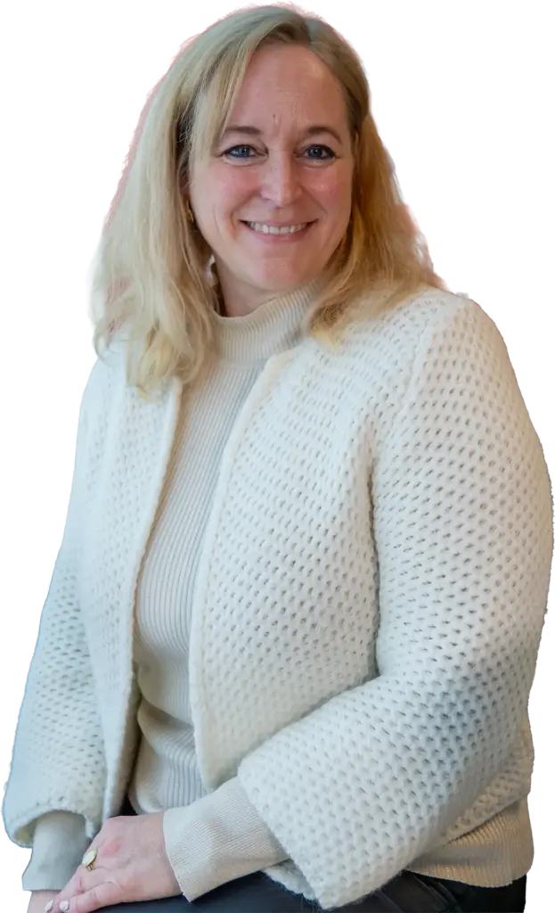 Lächelnde Frau mittleren Alters mit blonden schulterlangen Haaren, weißem Grobstrickcardigan und Rollkragenpullover, halbseitig sitzend.