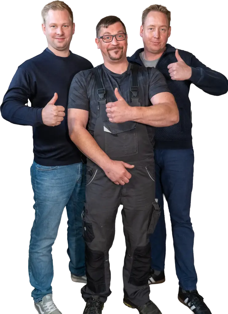 Drei lächelnde Männer, mittig ein Handwerker in Arbeitshose, alle mit Daumen hoch vor hellem, unscharf gestreiftem Hintergrund.