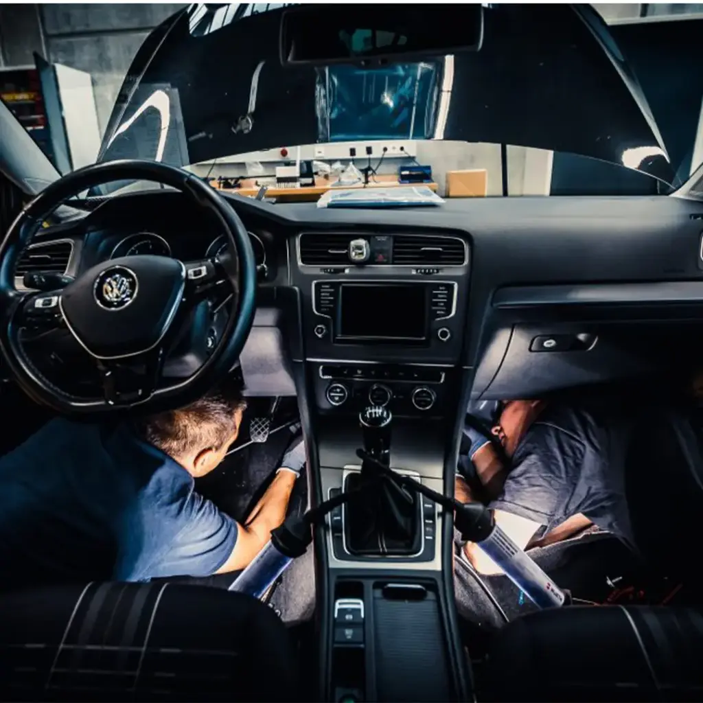 Zwei Mechaniker knien vorn im Innenraum eines VW, arbeiten unter Armaturenbrett und Pedalen bei geöffneter Motorhaube.