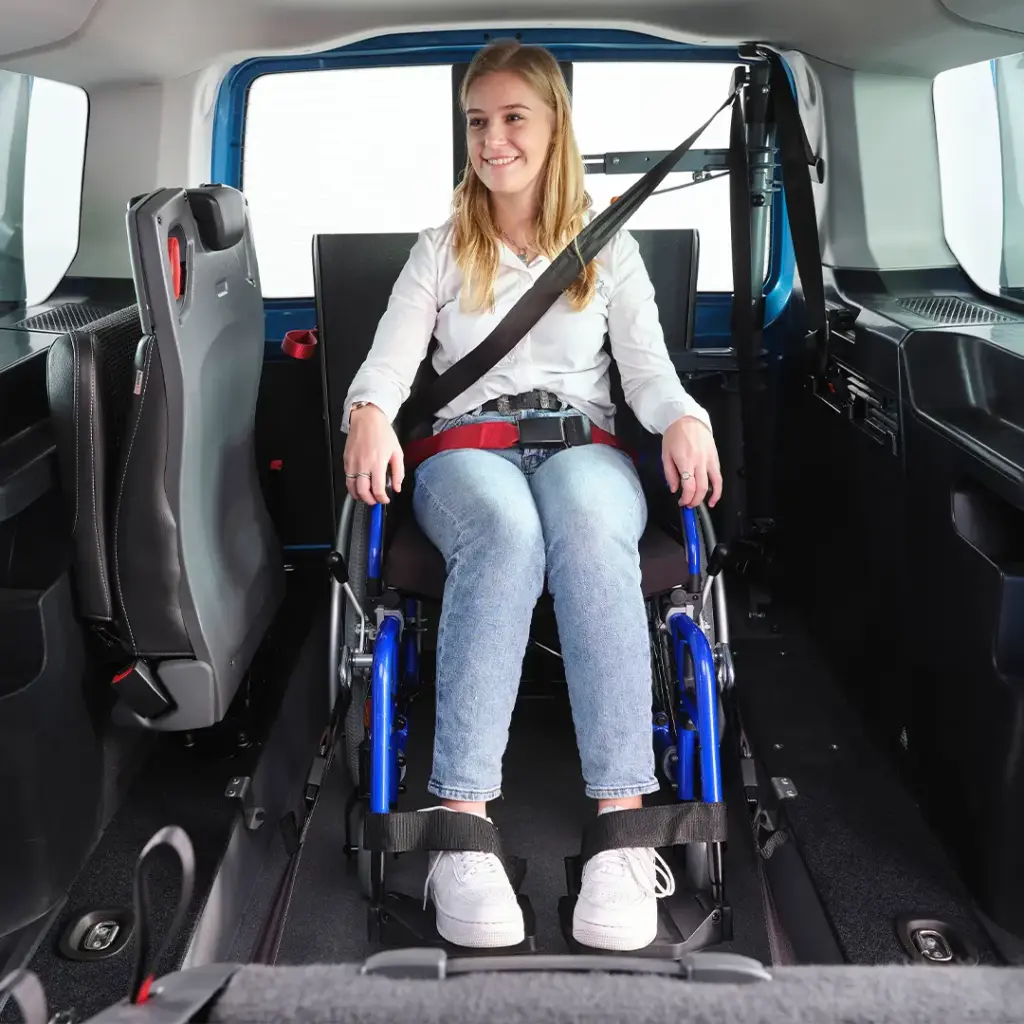 Junge Frau in einem Rollstuhl, angeschnallt mit Beckengurt und Schultergurt, sitzt im Innenraum eines Fahrzeugs und lächelt.