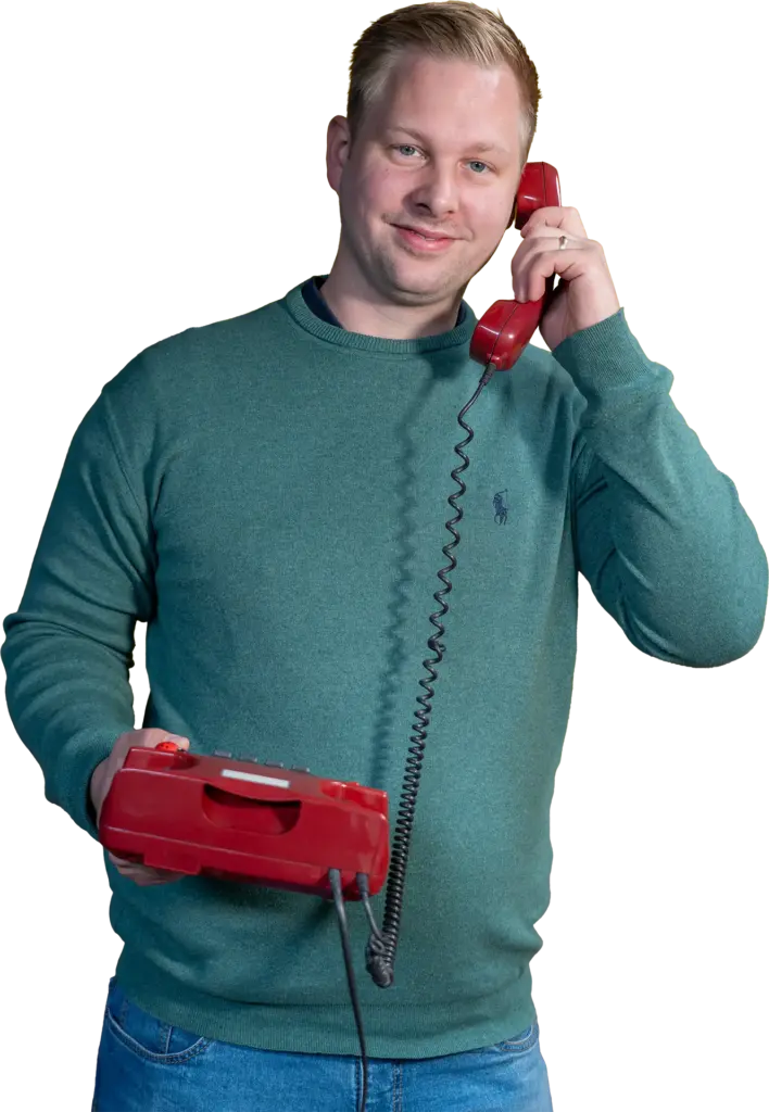 Mann mit grünem Pullover hält ein rotes Kabeltelefon ans Ohr und lächelt in die Kamera.