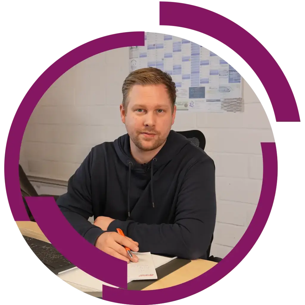 Mann in dunklem Hoodie sitzt am Schreibtisch, hält Stift über Notizblock, Bürowand mit Kalender im Hintergrund