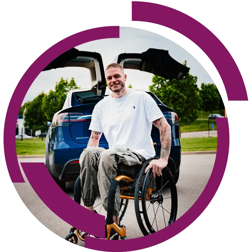 Mann im Rollstuhl mit Tattoos lächelnd vor offenem Heck eines blauen Autos, Parkumgebung im Hintergrund.