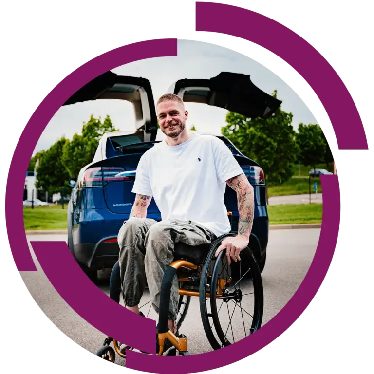 Mann im Rollstuhl mit Tattoos lächelnd vor offenem Heck eines blauen Autos, Parkumgebung im Hintergrund.