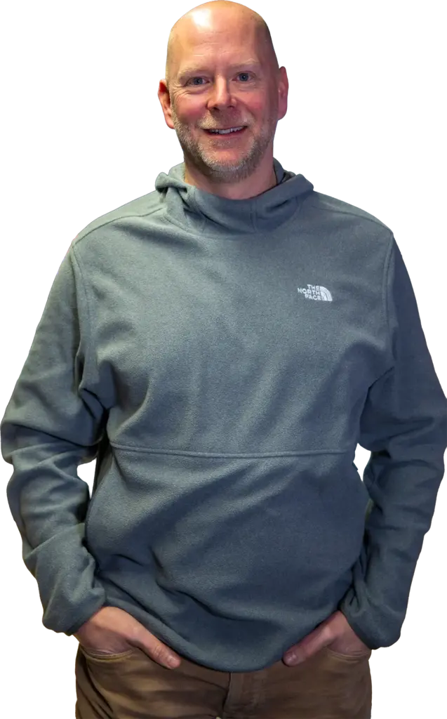 Bärtiger, glatzköpfiger Mann in grauem The North Face-Fleece mit Händen in den Hosentaschen und leichtem Lächeln.