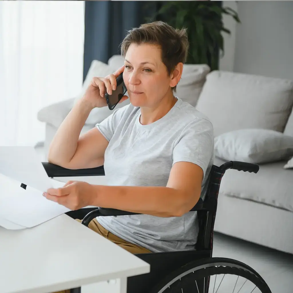 Frau im Rollstuhl am Tisch, hält Papiere und telefoniert mit Smartphone, ruhiges Wohnzimmer im Hintergrund.