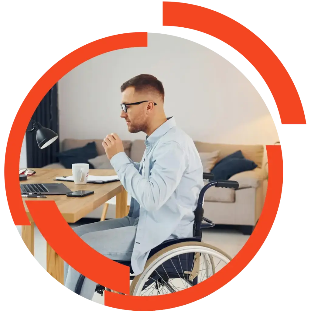 Mann im Rollstuhl arbeitet am Schreibtisch mit Laptop, Notizen und Kaffeetasse in einem hellen Wohnraum.