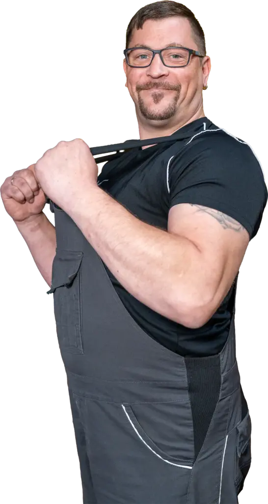 kräftiger Mann mit Brille, Schnurrbart und Tattoo trägt dunkle Arbeitslatzhose und zieht den Träger lächelnd über die Schulter