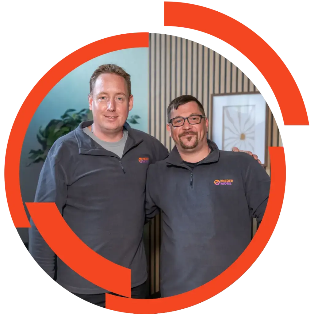 Zwei lächelnde Männer in grauen Firmenpullovern mit Logo, Arm in Arm vor einer gestreiften Wand und Pflanzen, roter Kreisrahmen um sie.