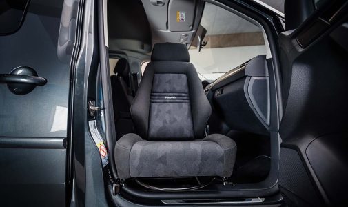 Geöffnete Vordertür eines Autos, im Vordergrund ein nach außen geschwenkter dunkelgrauer Recaro-Sitz mit dezentem Muster, im Hintergrund Armaturenbrett und Türrahmen.