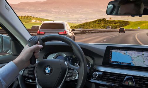 Blick aus dem Innenraum eines BMW: Hand am Lenkrad hält ein joystickähnliches Bediengerät, vor der Windschutzscheibe eine Autobahn mit mehreren Fahrzeugen und grünen Hügeln in der Ferne, rechts das Infotainment-Display mit Navigationskarte.