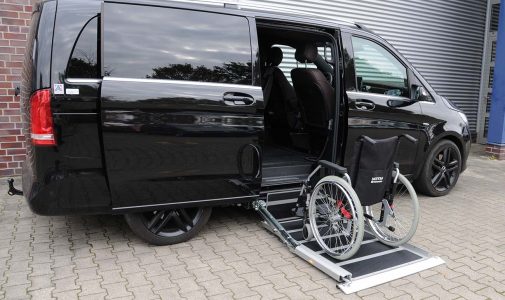 Schwarzer Van mit geöffneter Schiebetür und ausgefahrener Rampe, auf der ein Rollstuhl steht, geparkt vor einem Gebäude auf gepflastertem Hof.