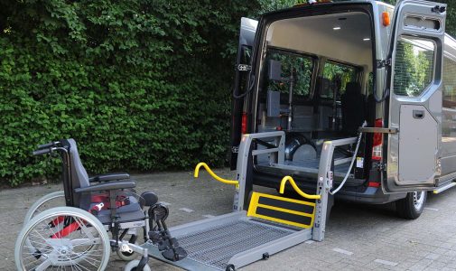 Manueller Rollstuhl steht neben einem offenstehenden silbernen Transporter, dessen ausgeklappte Metallrampe/Hubplattform mit markanten gelben Handläufen auf das gepflasterte Gelände führt; im Hintergrund eine dichte grüne Hecke.