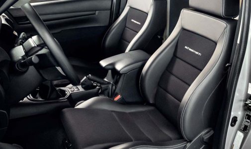 Innenraum eines Autos mit zwei schwarzen Sportsitzen mit heller Kontrastnaht und eingesticktem Schriftzug, mittlerer Armlehne, Schaltknüppel und Lenkrad sichtbar.