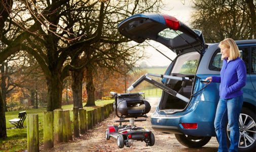 Frau steht neben dem geöffneten Kofferraum eines blauen Autos und bedient mit einer Fernbedienung einen Hebearm, der ein Elektromobil aus dem Kofferraum hebt, im parkähnlichen Umfeld mit Bäumen und Holzpfosten.